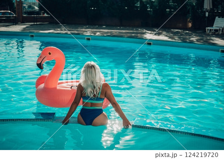 A woman in a bikini is sitting on a pink flamingo float in a pool. Concept of relaxation and leisure, as the woman enjoys her time in the water. 124219720