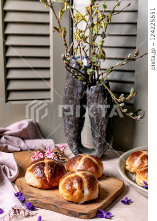 Hot cross buns on wooden cutting board with spring decoration. Easter pastry styled with willow branches in rustic vase, pink blossoms and violet flowers against vintage shutters background. 124225651