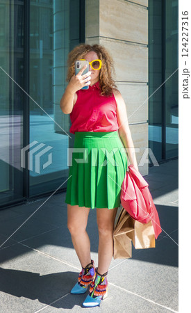 a beautiful young woman in bright clothes with a phone and bags takes selfies or takes pictures of the city on a walk 124227316