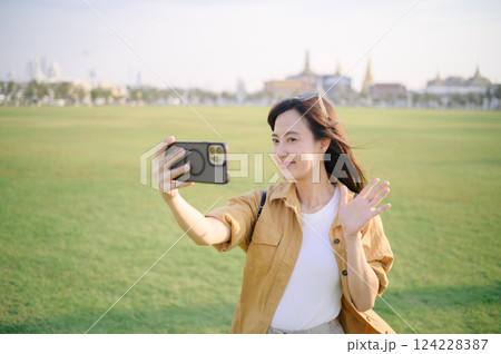 Traveler asian woman in her 30s making a livestream or selfie on smartphone while documents her unique Bangkok journey at Wat Pra Kaew. Connect with Buddhist tradition and discover the Emerald 124228387