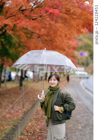 Asian woman in a stylish jacket smiles with an umbrella in the vibrant fall foliage. A delightful and cheerful portrait on a wet and colorful day. 124228451