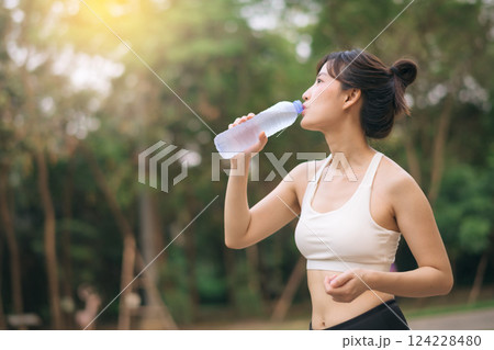 Female runner. Fit Asian young woman with white sportswear drinking water after running and enjoying a healthy outdoor. Fitness runner girl in public park. Wellness being concept Female runner. Fit Asian young woman with white sportswear drinking water after running and enjoying a healthy outdoor. Fitness runner girl in public park. Wellness being concept 124228480