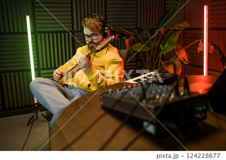 A focused male musician fine-tunes a guitar against the backdrop of a digital sound editing setup in a contemporary recording studio environment. 124228677
