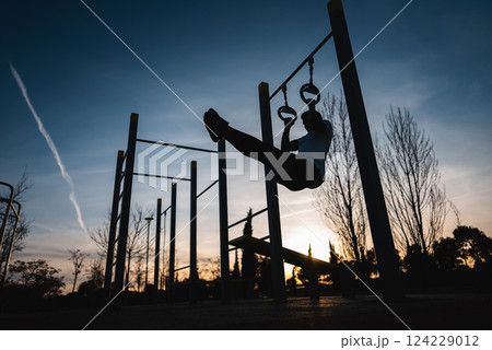young athletic man exercising fitness calisthenics workout and making ring leg raises for abdomen abs muscles at street gym during sunset, body improvement and strength training 124229012