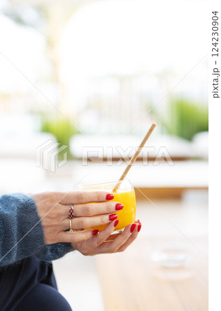 Close-up hands of woman holding glass of fresh orange juice, refreshing citrus drink for healthy lifestyle, minimalistic composition with vibrant blue and orange color and summer vibes 124230904