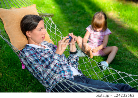 Mother relaxing in a hammock using a smartphone while her young daughter sits on the grass playing with her shoes in a sunny backyard, depicting family leisure and digital distraction outdoors 124232562