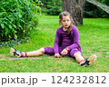 Young Girl in a Purple Outfit Sitting on Grass in a Green Garden, Looking Thoughtful and Curious 124232812