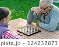 Elderly grandfather and young granddaughter playing checkers together at a wooden table in a garden, enjoying quality family time and strategic thinking outdoors in autumn 124232873