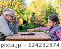 Elderly grandfather and young granddaughter playing checkers together at a wooden table in a garden, enjoying quality family time and strategic thinking outdoors in autumn 124232874