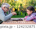 Elderly grandfather and young granddaughter playing checkers together at a wooden table in a garden, enjoying quality family time and strategic thinking outdoors in autumn 124232875