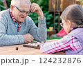 Elderly grandfather and young granddaughter playing checkers together at a wooden table in a garden, enjoying quality family time and strategic thinking outdoors in autumn 124232876
