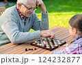 Elderly grandfather and young granddaughter playing checkers together at a wooden table in a garden, enjoying quality family time and strategic thinking outdoors in autumn 124232877
