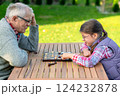 Elderly grandfather and young granddaughter playing checkers together at a wooden table in a garden, enjoying quality family time and strategic thinking outdoors in autumn 124232878