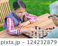 Young girl playing checkers with her grandfather at a wooden table outdoors, focusing on strategy and enjoying quality family time in a park or garden setting 124232879