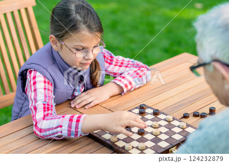 Young girl playing checkers with her grandfather at a wooden table outdoors, focusing on strategy and enjoying quality family time in a park or garden setting 124232879