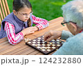 Young girl playing checkers with her grandfather at a wooden table outdoors, focusing on strategy and enjoying quality family time in a park or garden setting 124232880