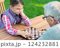 Young girl playing checkers with her grandfather at a wooden table outdoors, focusing on strategy and enjoying quality family time in a park or garden setting 124232881