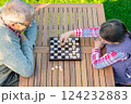 Elderly grandfather and young granddaughter playing checkers together at a wooden table in a garden, enjoying quality family time and strategic thinking outdoors in autumn 124232883