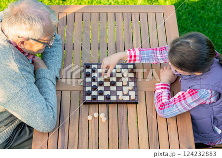 Elderly grandfather and young granddaughter playing checkers together at a wooden table in a garden, enjoying quality family time and strategic thinking outdoors in autumn 124232883