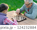 Elderly grandfather and young granddaughter playing checkers together at a wooden table in a garden, enjoying quality family time and strategic thinking outdoors in autumn 124232884