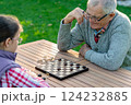 Elderly grandfather and young granddaughter playing checkers together at a wooden table in a garden, enjoying quality family time and strategic thinking outdoors in autumn 124232885