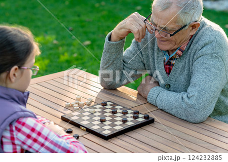 Elderly grandfather and young granddaughter playing checkers together at a wooden table in a garden, enjoying quality family time and strategic thinking outdoors in autumn 124232885