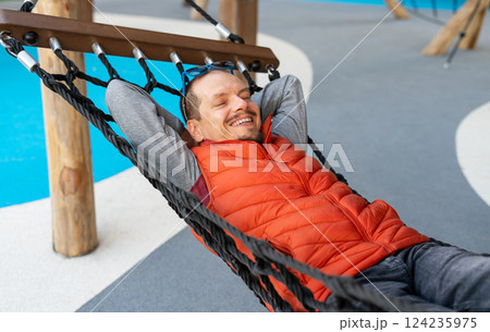 Relaxed man smiling contentedly while resting in hammock, wearing vibrant orange vest. His hands are behind his head, showcasing state of peacefulness and joy 124235975