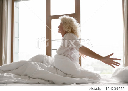 Awakened aged woman hotel guest sitting on bed stretching arms 124239632