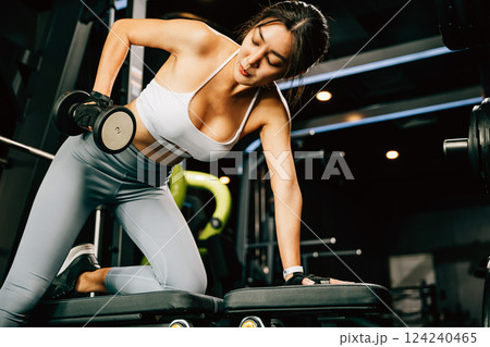 Asian woman lifting dumbbells on a bench in a fitness gym, happy young female performing strength training exercises with dumbbells in a gym, weight training for a healthy lifestyle concept 124240465