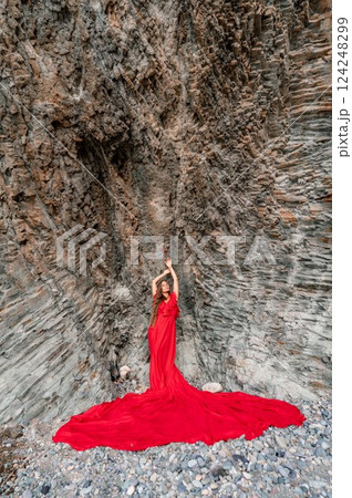 woman sea red dress. Woman with long hair on a sunny seashore in a red flowing dress, back view, silk fabric waving in the wind. Against the backdrop of the blue sky and mountains on the seashore. 124248299