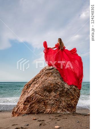 woman sea red dress. Woman with long hair on a sunny seashore in a red flowing dress, back view, silk fabric waving in the wind. Against the backdrop of the blue sky and mountains on the seashore. woman sea red dress. Woman with long hair on a sunny seashore in a red flowing dress, back view, silk fabric waving in the wind. Against the backdrop of the blue sky and mountains on the seashore. 124248301