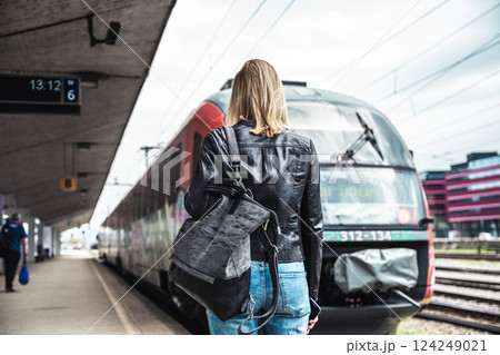 Young blond woman in jeans, shirt and leather jacket wearing bag and sunglass, embarking red modern speed train on train station platform. Travel and transportation 124249021
