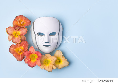 Cosmetic LED facial mask regenerative treatment on blue background with flowers lilies. Health and beauty of the face. Taking care of yourself and your face, anti-aging, prolonging youth and beauty 124250441