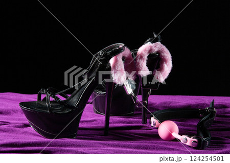 Adult sex games. Kinky lifestyle. Pink gag ball, handcuffs and a pair of black high-heeled shoes on the purple bedsheet. Bdsm outfit 124254501