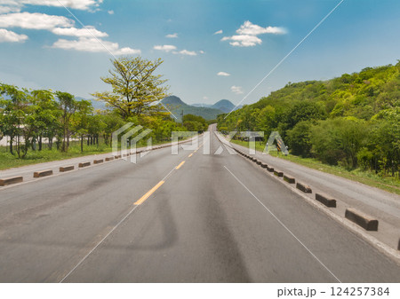 Asphalt road surrounded by greenery on a sunny day, Long asphalt road leading to a mountain with blue sky, Beautiful road on a sunny day leading to some mountains 124257384