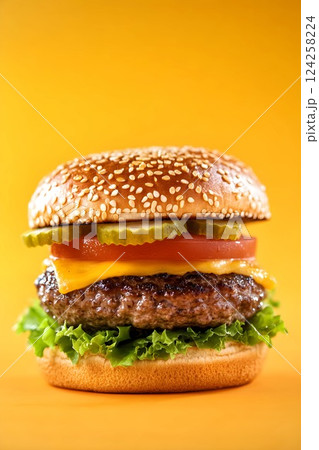 A cheeseburger consisting of a bun with sesame seeds, pickled cucumbers, tomatoes, cheese, meat patty and salad leaves on a yellow orange background 124258224