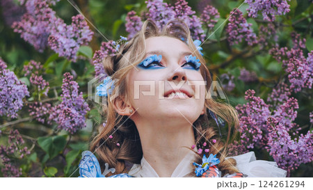 Female model sporting butterfly lashes, vibrant makeup, standing amid blooming lilac garden, inhaling springtime floral sweetness 124261294