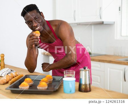 Cheerful naked man in red apron tasting muffin in home kitchen Cheerful naked man in red apron tasting muffin in home kitchen 124261316