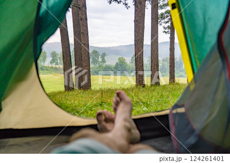 Camper relaxing inside tent, enjoying peaceful wilderness landscape with verdant meadow and dense forest backdrop 124261401