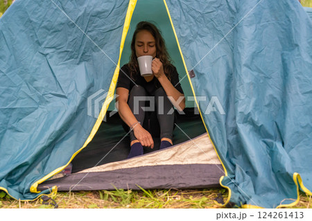Woman sipping steaming coffee, nestled inside rustic camping tent, morning sunlight filtering through pine trees surrounding wilderness campsite 124261413
