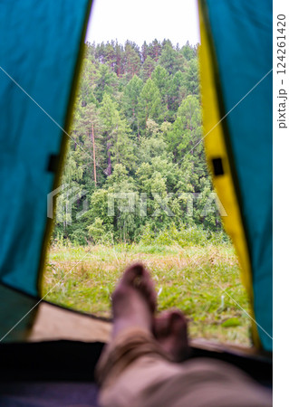 Legs of a camper relaxing inside a tent, soaking in the stunning view of a lush green forest and enjoying a moment of tranquility 124261420