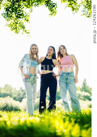 Female students smiling, standing close together in lush park setting, sharing summer friendship moment under bright sunlight 124261590