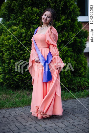 Graduate student wearing peach dress and graduation sash, beaming with joy amid blooming garden landscape Graduate student wearing peach dress and graduation sash, beaming with joy amid blooming garden landscape 124261592