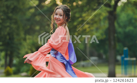 Female graduate rejoicing, spinning in pink dress with blue ribbon, park backdrop highlighting academic success and joyful transition 124261623