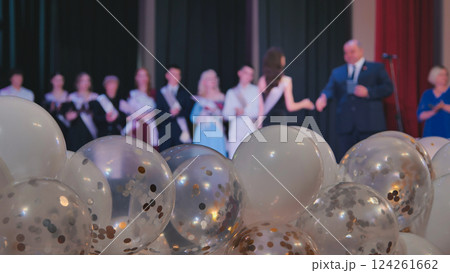 White and transparent balloons with gold and silver confetti fill the foreground, celebrating a high school graduation ceremony on stage 124261662