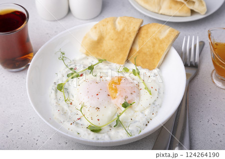 Turkish eggs (chilbir) with yogurt, fragrant butter, pea sprouts, flatbread and tea. Traditional Turkish breakfast with poached eggs (with liquid yolk). Selective focus, close-up. Turkish eggs (chilbir) with yogurt, fragrant butter, pea sprouts, flatbread and tea. Traditional Turkish breakfast with poached eggs (with liquid yolk). Selective focus, close-up. 124264190