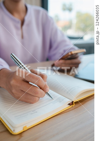Close-up of a woman writing in a notebook while holding a smartphone, set in a bright office space. 124266035