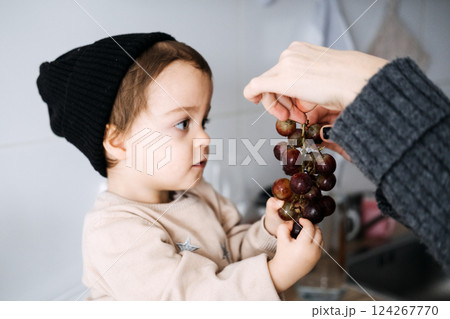 Mother offering toddler grapes, encouraging healthy eating habits. Toddler nutrition, early childhood development, family care, nurturing moments, healthy diet. 124267770