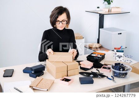 Female entrepreneur packing sustainable cardboard boxes for her handmade business. Eco-friendly packaging, sustainable e-commerce, ethical branding, small business, conscious consumerism. Female entrepreneur packing sustainable cardboard boxes for her handmade business. Eco-friendly packaging, sustainable e-commerce, ethical branding, small business, conscious consumerism. 124267779