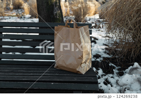 A paper craft bag with Takeaway food products groceries stands on a wooden bench in a winter sunny day. Fast delivery 124269238
